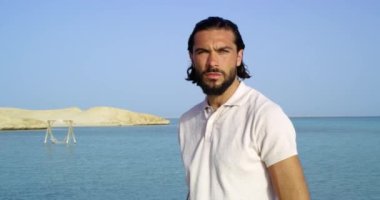 Sabit çekim, kahverengi tenli, uzun saçlı, gözlerinde keskin bir bakış olan yakışıklı bir adam, plajda duran beyaz gömlekli bir adam ve güneşli bir günde arkasında kızıl denizi izleyen bir arka plan..