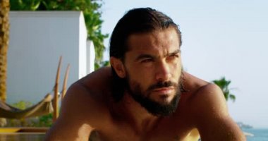 Yakın plan çekimde adamın yüzü kahverengimsi tenli, adamın kestane rengi gözleri ve kaşları çatık, kalın koyu renk sakalı ve uzun saçları var, yazın havuz kenarında oturan atletik vücutlu bir adam..