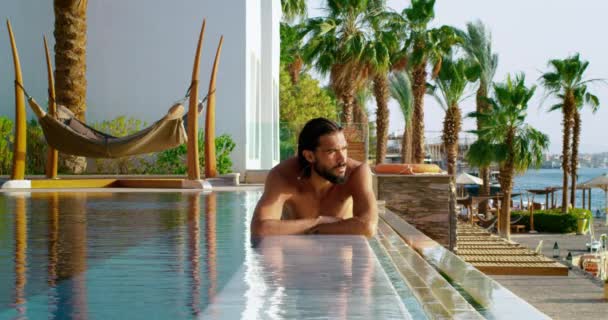 涼しいさわやかな水で泳いだ後 スイミングプールの端に彼の胃の上に横たわる半裸の日焼けした皮膚の男 素晴らしい景色とカラフルな背景を持つエジプトの豪華なホテルのプール — ストック動画