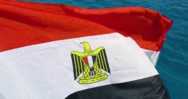 Rüzgarda dalgalanan Mısır bayrağının aşırı yakın çekimi. Kızıl denizin suları arkasında beliriyor. Çekim, ortasında kartal sembolü olan üç yatay kırmızı, beyaz ve siyah şeritten oluşan üç renkli bayrağı gösteriyor.