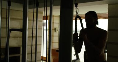 Spor salonunda çalışan bir adam erkeklik organını kaldırıyor. Güzel göğüslü adam spor salonunda antrenman yapıyor, ağırlık kaldırıyor ve kaslı aletler kullanıyor, açık bir pencereden gökyüzünün arka planına bakan karanlık bir sahne.