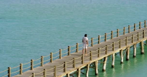 海の中の木製の橋を歩いて男の高角度ショット 夏の日に海の透明度の高い水の美しい景色と海の真ん中の木製のデッキを歩いて後ろから男の高い角度のショット — ストック動画