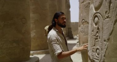 Gezgin, antik Mısır bölgelerindeki bir sütuna oyulmuş eski işaretlere yakından bakıyor, bazı işaretler çevresel elementler nedeniyle yok edilmiş, sütunlar büyük bir kum taşı kütlesinden yapılmış.