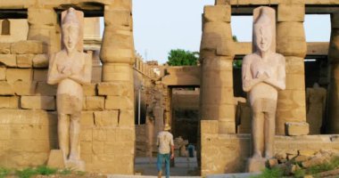 Antik bir tarihi kilisenin girişinde duran iki büyük Mısırlı muhafız heykeli, dev kapıdan geçen bir erkek turist, büyük sarı taşlarla inşa edilmiş, tapınağın ön cephesinde birçok pencere var.,