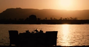Nehir kenarında romantik bir masa, güneşin dağların ardında kaybolduğu sihirli bir saatte mükemmel bir manzara, güneş ışığı nehri sihirli bir altın renkle boyuyor, masanın yanındaki çift için iki sandalye hazır.