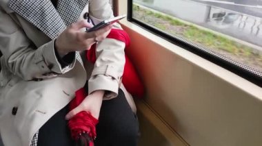 Slow motion. A young girl in a beige raincoat with a bag and a red umbrella rides the bus and writes a message on a mobile phone.