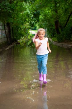 A happy braided girl in rubber boots is jumping and running in muddy puddles.