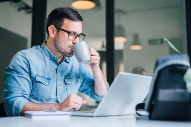 Ev bilgisayarında bilgisayar başında çalışan, kahve içen, sıradan girişimci küçük işletme sahibi, elinde kahve fincanı tutarak araştırma yapan yakışıklı bir adamın portresi.