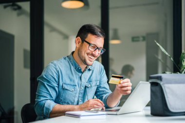 Mutlu, gülümseyen, yetişkin bir adam online alışveriş yapıyor ya da online ödeme yapan girişimci ya da serbest çalışan serbest çalışan mutlu ücret alıyor.