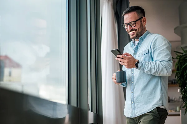 Uśmiechnięty Biznesmen Dorywczo Stojący Biurze Przy Oknie Przy Użyciu Telefonu — Zdjęcie stockowe