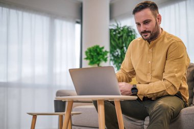Odaklanmış iş adamı serbest girişimci evinde rahat bir kanepede otururken ev bilgisayarında bilgisayarla çalışıyor.