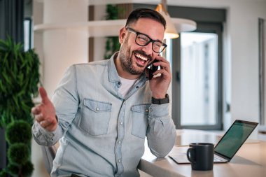 Deneyimli bir iş adamı telefonda gülümsüyor. Mutlu, genç, yetişkin iş adamı iş ortaklarıyla telefonda iletişim kuruyor. Modern bir çalışma alanında başarılı bir girişimci.
