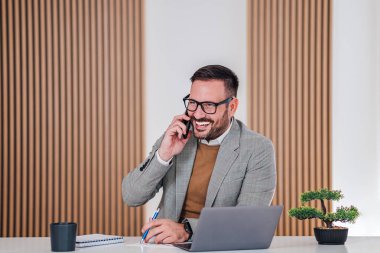 İş adamı iş yerindeki akıllı telefon anlaşmalarını kullanarak cep telefonuyla konuşan mutlu, genç, yetişkin bir işadamı arıyor.