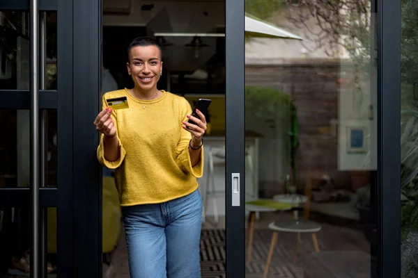 カメラを見てクレジットカードを使用してドアのフレームにもたれて自宅からオンラインショッピングの目の前でオンライン決済のための電話を使用して笑顔の女性の肖像明るく幸せな — ストック写真