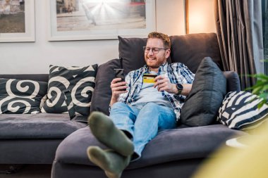 Neşeli yetişkin bir adam kredi kartı ve akıllı telefonla evinin kanepesinde rahatça oturup online ödeme alışverişi yapıyor ve online bankacılık yapıyor.