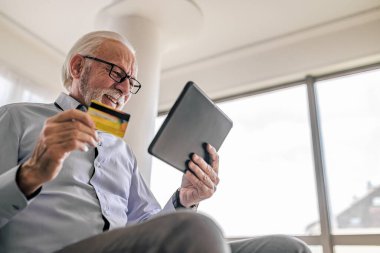 Beyaz saçlı ve sakallı yaşlı bir adamın online alışveriş için dizüstü bilgisayar ve kredi kartı kullanmasının düşük açılı görüntüsü veya internet ödemesi