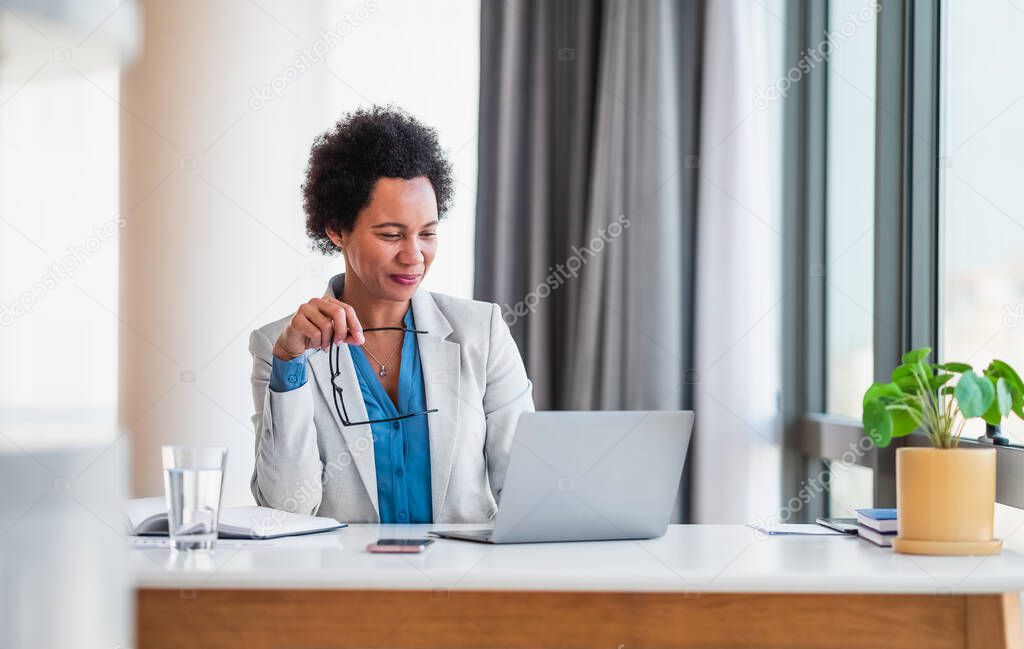 Smiling young adult mature African American businesswoman working on a laptop at her desk in a bright modern office holding her glasses.