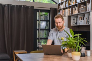 Ciddi bir yetişkin adam, ev ofisinde, bilgisayarında çalışıyor..