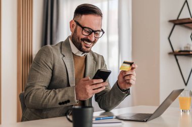 Akıllı telefonla kart ödemesi yapan kendine güvenen bir işadamı. Mutlu profesyoneller çevrimiçi alışveriş yapıyor. Ofis masasında dizüstü bilgisayarla çalışıyor..