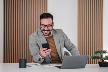Gülümsemek, cep telefonundaki profesyonel iş mesajlarının dikkatini dağıtıyor. Dizüstü bilgisayarda çalışan mutlu girişimci. Ofisteki masada oturuyor..
