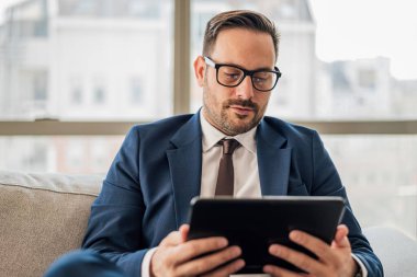 Dijital tablet kullanan kendine güvenen bir iş adamı. Erkek profesyonel takım elbise giyiyor. Ofisteki koltukta otururken kablosuz bilgisayar kullanıyor..