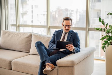 Ofiste dijital tablet kullanan kendinden emin bir erkek girişimci. İş adamı pencereye karşı kanepede oturuyor. İş yerindeki kablosuz bilgisayarda çalışıyor..