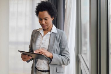 Dijital tablet kullanan kendine güvenen bir iş kadını. Kadın girişimci pencerenin önünde duruyor. O, ofiste çalışırken forma giyiyor..