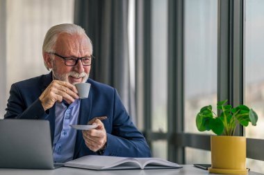 Gülümseyen yaşlı profesyonel kahve içiyor. Üst düzey işadamı masada dizüstü bilgisayarla çalışıyor. Ofiste formalite ve gözlük takıyor..