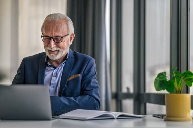 Dizüstü bilgisayarda çalışan gülümseyen yaşlı iş adamı. Taşınabilir cihaz kullanırken format giyen son sınıf erkek profesyonel. Takım elbiseli, ofiste masa başında oturuyor..