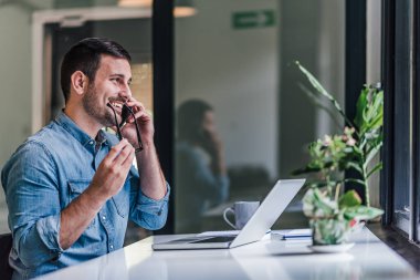 Gözlüklü, akıllı telefonla konuşan, gülümseyen, serbest çalışan bir erkek. İş adamı masada dizüstü bilgisayar kullanıyor. Ofiste çalışıyor..