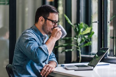 Dizüstü bilgisayar kullanırken kahve içen genç bir serbest yazar. İş adamı gündelik kıyafet ve gözlük takıyor. Ofisteki masada çalışıyor..