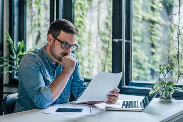 Serious entrepreneur analyzing documents. Male professional reading reports while working on laptop. He is sitting at table in home office.