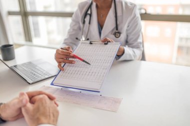 Kadın doktor erkek hastaya sağlık raporunu açıklıyor, sağlık görevlisi masadaki adamla tartışıyor, hastanenin penceresinde oturuyorlar..