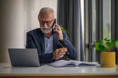 Kendine güvenen kıdemli işadamı cep telefonuyla konuşuyor, erkek profesyonel masa başında dizüstü bilgisayarda çalışıyor, ofis masasında otururken birden fazla iş yapıyor..