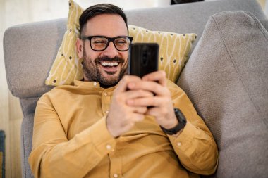 Neşeli genç adam akıllı telefondan video izliyor, mutlu erkek oturma odasında kanepede dinleniyor, evde gözlük takıyor..