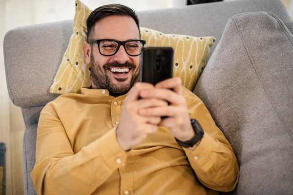 Neşeli genç adam akıllı telefondan video izliyor, mutlu erkek oturma odasında kanepede dinleniyor, evde gözlük takıyor..
