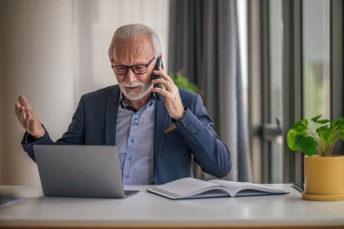 Gergin iş adamı dizüstü bilgisayarla çalışırken cep telefonuyla konuşuyor. Son sınıf erkek profesyonelin forma giymesinden endişe ediyorum. Şirket ofisindeki masada oturuyor..