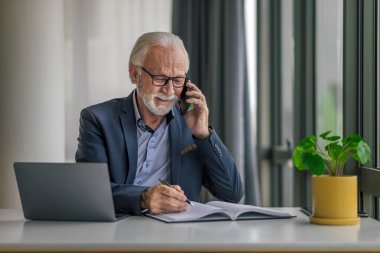 Kendine güvenen kıdemli işadamı cep telefonuyla konuşuyor. Erkek profesyonel masadaki dizüstü bilgisayarda çalışıyor. İşletme masasında otururken birden fazla iş yapıyor..