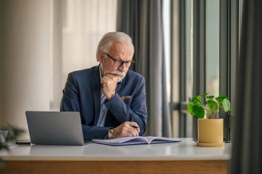 Kendine güvenen kıdemli işadamı notlar alıyor, düşünüyor ve planlıyor. Erkek profesyonel masadaki dizüstü bilgisayarda çalışıyor. Ofis masasında otururken birden fazla iş yapıyor..