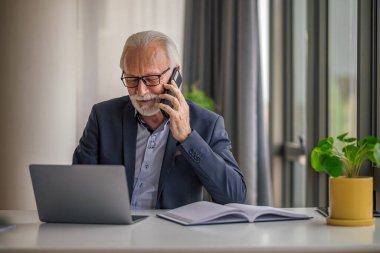 Kendine güvenen kıdemli işadamı cep telefonuyla konuşuyor, erkek profesyonel masa başında dizüstü bilgisayarda çalışıyor, ofis masasında otururken birden fazla iş yapıyor..