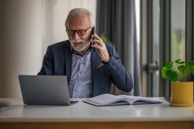Gergin iş adamı dizüstü bilgisayarla çalışırken cep telefonuyla konuşuyor. Son sınıf erkek profesyonelin forma giymesinden endişe ediyorum. Şirket ofisindeki masada oturuyor..