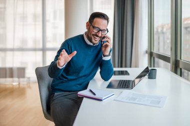 Akıllı telefonda konuşurken neşeli iş adamı el kol hareketi yapıyor. Erkek profesyonel iş kıyafetleri giyiyor. Şirket işyerindeki masada oturuyor..