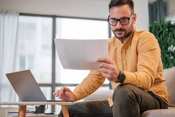 Belgeleri analiz eden ciddi bir serbest çalışan erkek. Dizüstü bilgisayar üzerinde çalışırken erkek profesyonel okuma kağıtları. Ofisteki kanepede oturuyor..