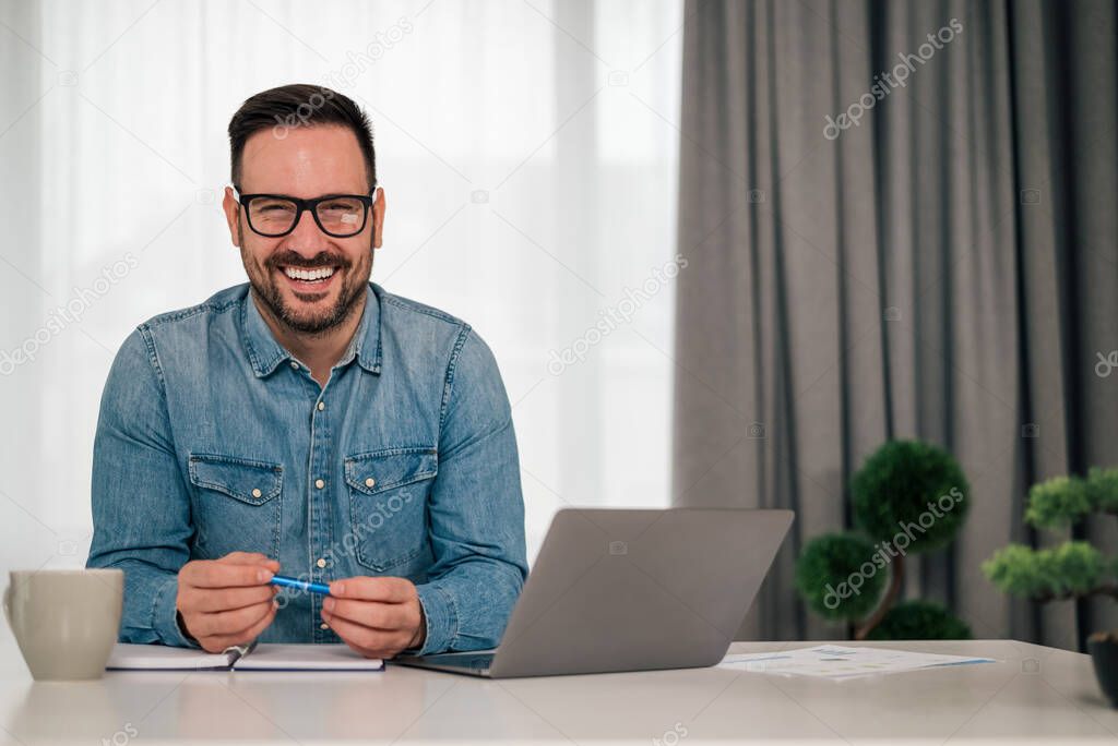Portrait of happy successful businessman with laptop and diary. Young entrepreneur is wearing eyeglasses. He is sitting at desk in office.