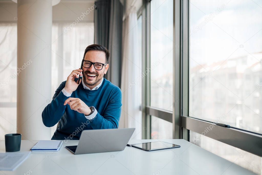Portrait of happy business professional discussing on mobile phone. Young smiling businessman is wearing smart casuals. He is sitting at desk in corporate office.