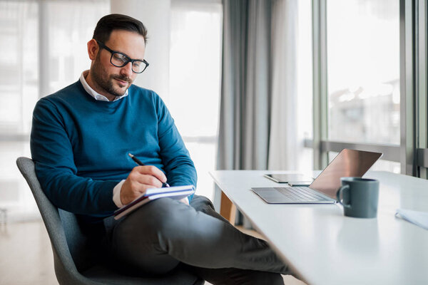 Confident entrepreneur writing business plan in diary. Young professional is planning strategy. He is working while sitting at desk in office.