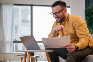 Belge okuyan, gülümseyen genç bir serbest yazar. İş adamı dizüstü bilgisayarla çalışırken iş planını analiz ediyor. Ofisteki kanepede oturuyor..