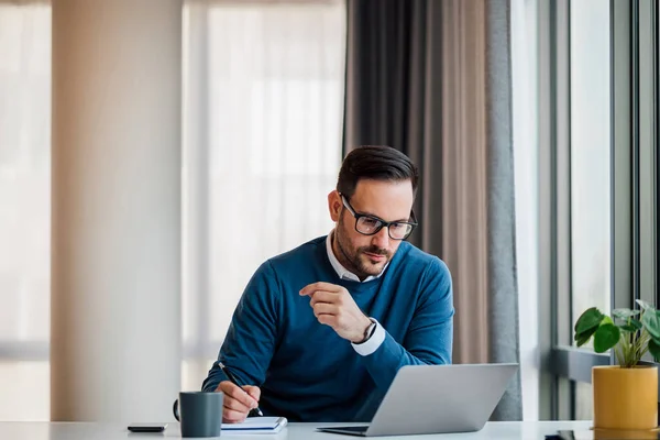 Confident business professional making growth plan in diary. Male entrepreneur is working on laptop at desk. He is wearing smart casuals in workplace.