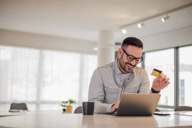 Neşeli girişimci kredi kartıyla online ödeme yapıyor. Masada dizüstü bilgisayarda çalışan genç bir iş adamı. Ofiste şık kıyafetler giyiyor..