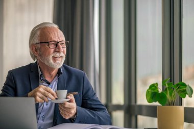 Üst düzey işadamı dizüstü bilgisayarla çalışırken kahve içiyor. Yaşlı profesyonel erkek başka tarafa bakıyor. Ofiste gözlük takıyor..